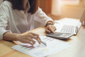 A imagem exibe uma mulher, uma calculadora, um notebook e alguns papéis em cima de uma mesa, representando a organização de um fluxo de caixa.